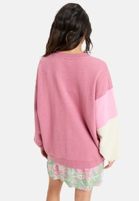 Lyserød oversized sweater med et lyserødt og cremefarvet farveblokdesign, der har en ribbet halsudskæring og struktureret stof, parret med en blomsterkjole.