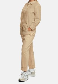 Camicia beige a maniche lunghe con colletto e davanti bottonato, abbinata a pantaloni beige con risvolto e scarpe da ginnastica bianche con dettagli grigi.