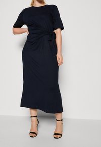 Marineblauwe, korte mouwen maxi-jurk met een gestrikt taille-detail, gemaakt van soepele stof. Gecombineerd met zwarte hakken met bandjes, tegen een effen achtergrond.