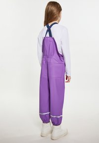 Violetinės vandeniui atsparios kombinezono kelnais su tekstūruotu paviršiumi, su elastinėmis rankogaliais ir reguliuojamomis mėlynomis petnešomis, derinamos su baltomis botais.