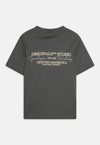 Grå bomull t-skjorte med korte ermer. På baksiden er det trykket tekst i hvitt og lys beige, inkludert "ORIGINALS™ STUDIO" og "Crafted Aesthetics."