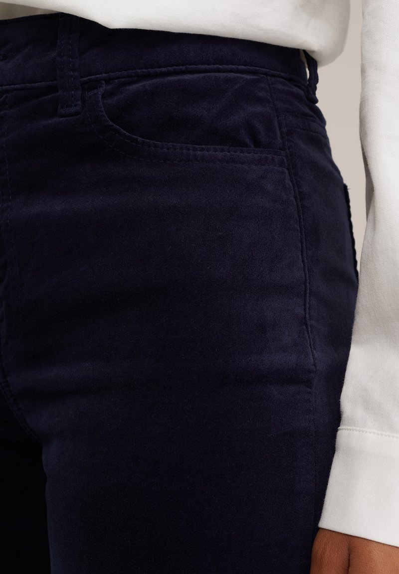 Pantalones de pana azul marino con una textura suave, que presentan un bolsillo frontal y presillas para cinturón. Camisa blanca de manga larga parcialmente visible.