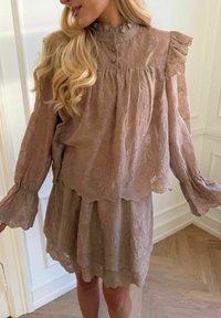 Femeie purtând o rochie bej cu dantelă, cu margini zimțate, detalii cu volane la umeri, mâneci lungi și ample și nasturi lângă un gât înalt.