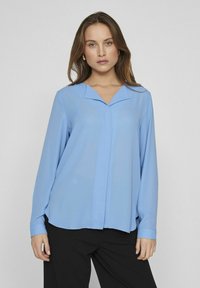 Lyseblå langermet bluse med V-formet krage, jevn tekstur og knappedetalj foran. Kombinert med svarte bukser.