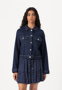 Mørk denim kuttet jakke med knappelukking, to brystlommer og spredt krage, paret med en marineblå prikkete kjole med volanger.