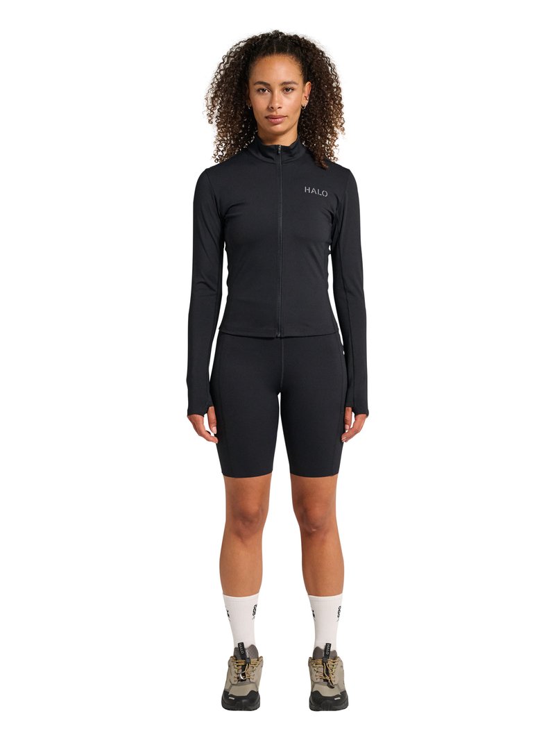 Kvinde med krøllet hår iført en sort langærmet trøje med lynlås mærket "HALO,", sorte cykelshorts, hvide sokker og beige vandresko.