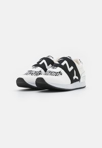 Weiße und schwarze Sneaker mit einer strukturierten Oberfläche, ausgestattet mit einem auffälligen schwarzen Riemen, geometrischen Mustern und einer gepolsterten Sohle.