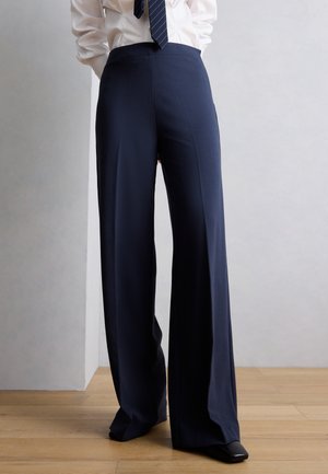 Persoană purtând pantaloni largi bleumarin, cămașă albă și cravată în dungi, stând pe podea de lemn în fața unui perete simplu, luminos.