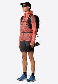 Lekki, pomarańczowy, wodoodporny kurtka z czarnymi akcentami, w połączeniu z ciemnymi szortami i niebieskimi butami trekkingowymi; trzymając kije trekkingowe.