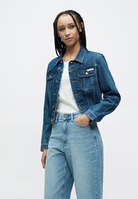 Tumesinise teksajope kahete esi taskuga, hõbedaste nööpide ja rinnal sildiga, kombineeritud helesiniste kõrge vöökohaga teksapükstega.