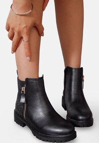 Bottines noires en cuir lisse avec un panneau latéral texturé, fermeture éclair et semelle en caoutchouc, présentées sur un pied avec une main touchant la cheville.