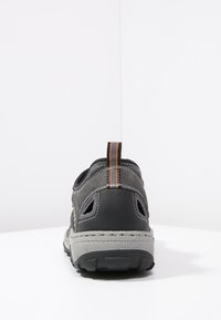 Chaussure de sport noire et grise avec des panneaux en mesh, une semelle en caoutchouc texturée et une bande d'accent orange sur le tirant du talon.