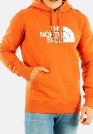 Muž nosící oranžovou mikinu The North Face s přední kapsou a světle modré džíny, ruka částečně v kapse.