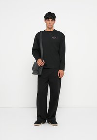 Schwarz langärmliges Hemd mit pinkem Logo, kombiniert mit lockeren schwarzen Hosen und schwarzen Espadrilles. Eine schwarze Schultertasche rundet den Look ab.