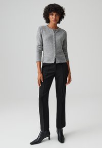 Cardigan grigio lavorato a maglia con scollo rotondo e chiusura a bottoni, abbinato a pantaloni neri su misura e stivaletti neri con tacco.