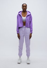 Κοντή μωβ φούτερ με κουκούλα πάνω από γκρι σπορ μπουστάκι, συνδυασμένη με μωβ παντελόνια φόρμας με ελαστική μέση και μανσέτες. Λευκά αθλητικά παπούτσια.