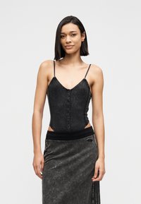 Νεαρή γυναίκα με ίσια σκοτεινά μαλλιά, ντυμένη με μαύρο αμάνικο τοπ με κουμπιά και σκουρόχρωμη γκρι φούστα, μπροστά σε απλό φωτεινό φόντο.