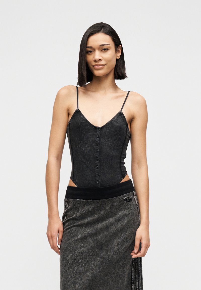 Νεαρή γυναίκα με ίσια σκοτεινά μαλλιά, ντυμένη με μαύρο αμάνικο τοπ με κουμπιά και σκουρόχρωμη γκρι φούστα, μπροστά σε απλό φωτεινό φόντο.