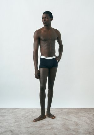 Pikk, kõhn mees tumeda nahaga, mustades Nike bokserites, paljajalu vaibal seistes, vaatamas küljele heledal ühtlasel taustal.