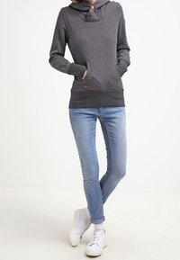 Sudadera gris con cuello tipo cowl y detalle de botones, con un patrón de puntos y bolsillos frontales, combinada con jeans de color azul claro y zapatillas blancas.