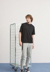 T-shirt nera a maniche corte abbinata a pantaloni della tuta grigi chiari con logo. Sneakers bianco e nere. Stendino in metallo sullo sfondo.