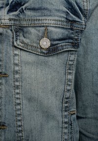 Ljusblå denimjacka med synliga sömmar, en metallknapp med logotyp och små fickdetaljer på bröstet. Slät, klassisk design.