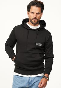 Schwarzer Hoodie mit einer vorderen Tasche, Kordelzugkapuze und weißen Logodetails auf der Brust, kombiniert mit hellblauen Jeans.