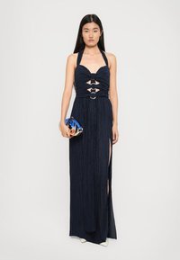 Navy halter maxi dress with pleated texture, front cutouts, and metal accents. Model holds a round blue clutch. High slit on one side.