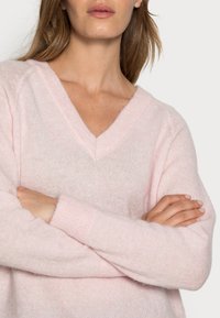 Kvinde med krydsede arme iført en blød lyserød V-hals sweater, vist fra skuldre til øvre bryst, neutral baggrund.