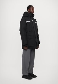 Crna oversized parka jakna s reflektirajućim trakama, više džepova i velikom kapuljačom; kombinirana s sivoj hlačama širokog kroja i crnim cipelama.