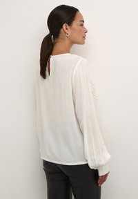 Blouse blanche avec une ouverture en forme de clé au dos, manches longues bouffantes et détails brodés délicats. Tissu lisse et coupe décontractée. Associée à un pantalon noir.