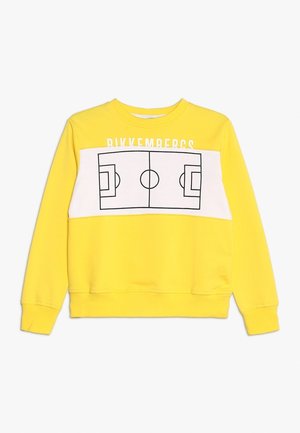 Żółta bluza z białym poziomym panelem z nadrukiem czarnego konturu boiska do piłki nożnej oraz napisem „BIKKEMBERGS” nad nim.