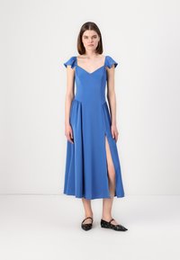 Robe maxi bleue avec un décolleté en V et des manches courtes à volants, dotée d'une fente sur le côté et d'une jupe fluide. Portée avec des ballerines noires.