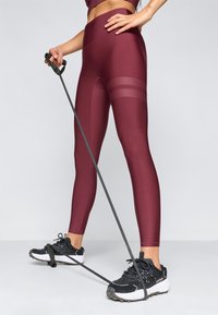 Bordowe legginsy z wysokim stanem charakteryzują się gładką teksturą i wzorzystymi akcentami. Na nogach są czarne buty sportowe z białymi podeszwami i sznurówkami.