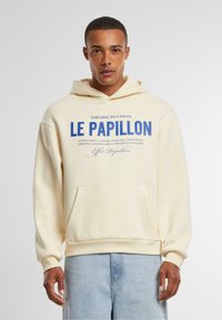 Crème hoodie met een buidelzak, voorzien van gedurfde blauwe tekst. Losse fit met ribgebreide manchetten en onderrand. Zachte textuur, eenvoudig ontwerp.