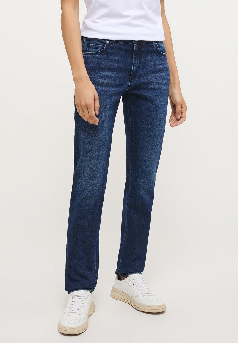 Osoba oblečená v tmavě modrých džínách, bílých teniskách a bílém tričku s krátkým rukávem, stojící na jednobarevném pozadí.