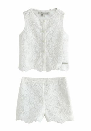 Mädchen mit langen Haaren trägt ein weißes ärmelloses Spitzentop und passende Shorts, steht mit überkreuzten Beinen und schaut seitlich, trägt goldene Sandalen.