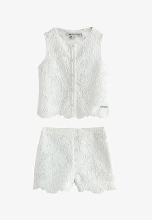 Dziewczyna z długimi włosami nosi biały koronkowy top bez rękawów i pasujące szorty, stoi ze skrzyżowanymi nogami i patrzy na bok, mając na nogach złote sandały.