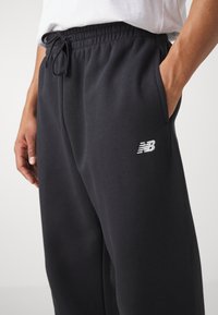 Schwarze Baumwoll-Sweatpants mit Kordelzugbund, Seitentaschen und einem kleinen weißen New Balance-Logo am linken Bein. Weicher Stoff.