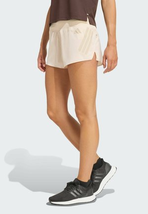 Osoba w beżowych spodenkach do biegania Adidas i czarnych butach sportowych Adidas, stojąca na tle jasnego tła.