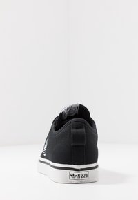 Černá plátěná teniska s bílou podrážkou, představující plochý design, texturovaný svršek a bílé logo Adidas Nizza na patě.