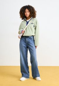 Fleecepullover i ljusgrön med en blå krage och logotyp, i kombination med vida blå jeans och vita sneakers; modellen poserar mot ett gult golv.