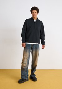 Sudadera negra de medio cierre, jeans de mezclilla con un diseño desgastado, parches en las rodillas y botas oscuras. Ambientado contra un fondo claro con un suelo amarillo.