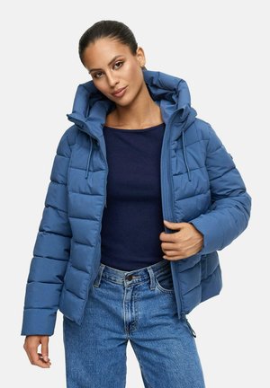 Femme portant une doudoune matelassée bleue sur un haut bleu foncé et un jean bleu, les cheveux tirés en arrière et une main ajustant la fermeture éclair de la veste.