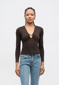 Haut marron à manches longues avec un décolleté en V profond, présentant un tissu froncé et un détail de boucle métallique au centre. Associé à un jean bleu clair.