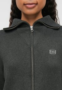 Mørkegrå jakke med glidelås i ribbestrikket tekstur, har krage, sølvglidelås og en liten logo på venstre brystområde.