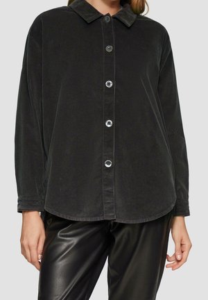 Woman wearing a black corduroy button-up shirt with long sleeves and black leather pants against a plain background.