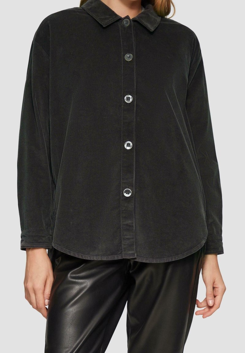 Femme portant une chemise en velours côtelé noire à boutons avec manches longues et un pantalon en cuir noir sur un fond uni.
