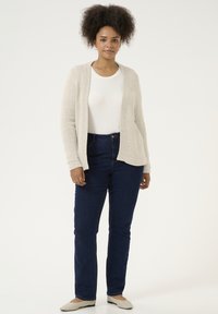 Cardigan beige clair avec une ouverture sur le devant, tricot texturé, porté sur un haut blanc, associé à un jean bleu foncé et des ballerines claires.