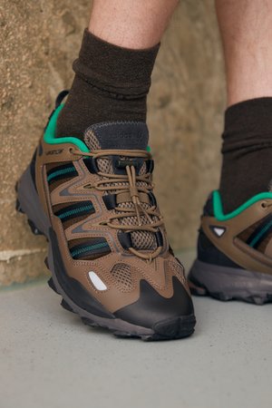 Άτομο που φορά καφέ και μαύρα ορειβατικά παπούτσια με πράσινη επένδυση και καφέ κάλτσες, στέκεται σε μια γκρίζα επιφάνεια κοντά σε μια υφασμένη τοιχοποιία.